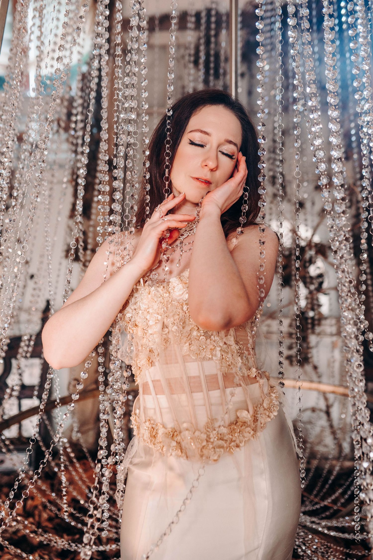Elegant woman in gold embellished corset gown poses gracefully among hanging crystal bead curtains in a luxurious setting.