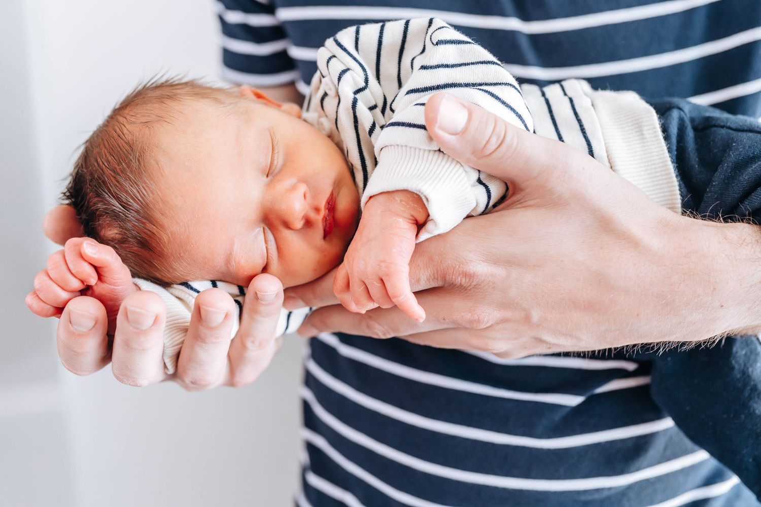 Newborn baby sleeping peacefully in parent's hands, wearing white striped onesie, close-up portrait photo.