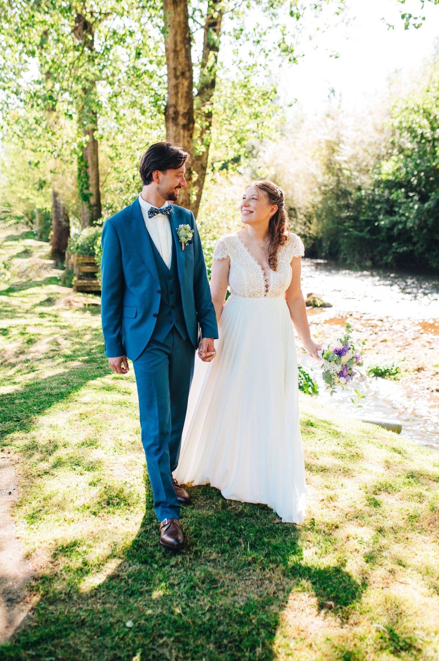 Couple in wedding attire walking through sunlit garden with trees.