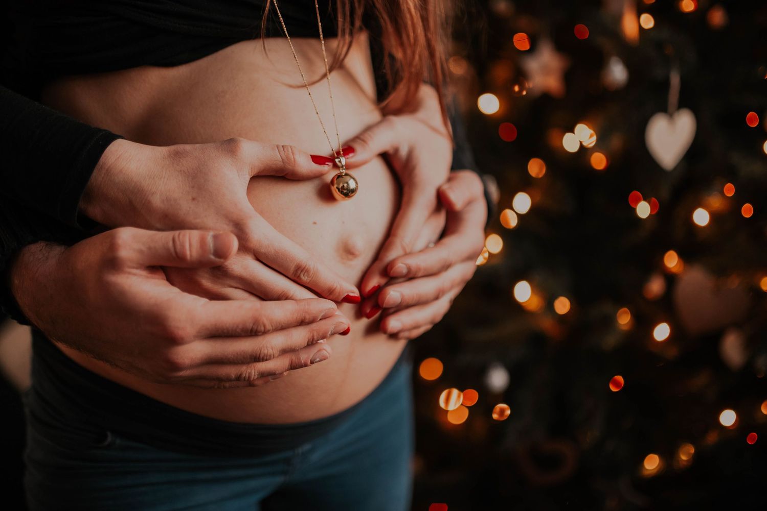 Hands form heart shape on pregnant belly with Christmas lights twinkling in background.