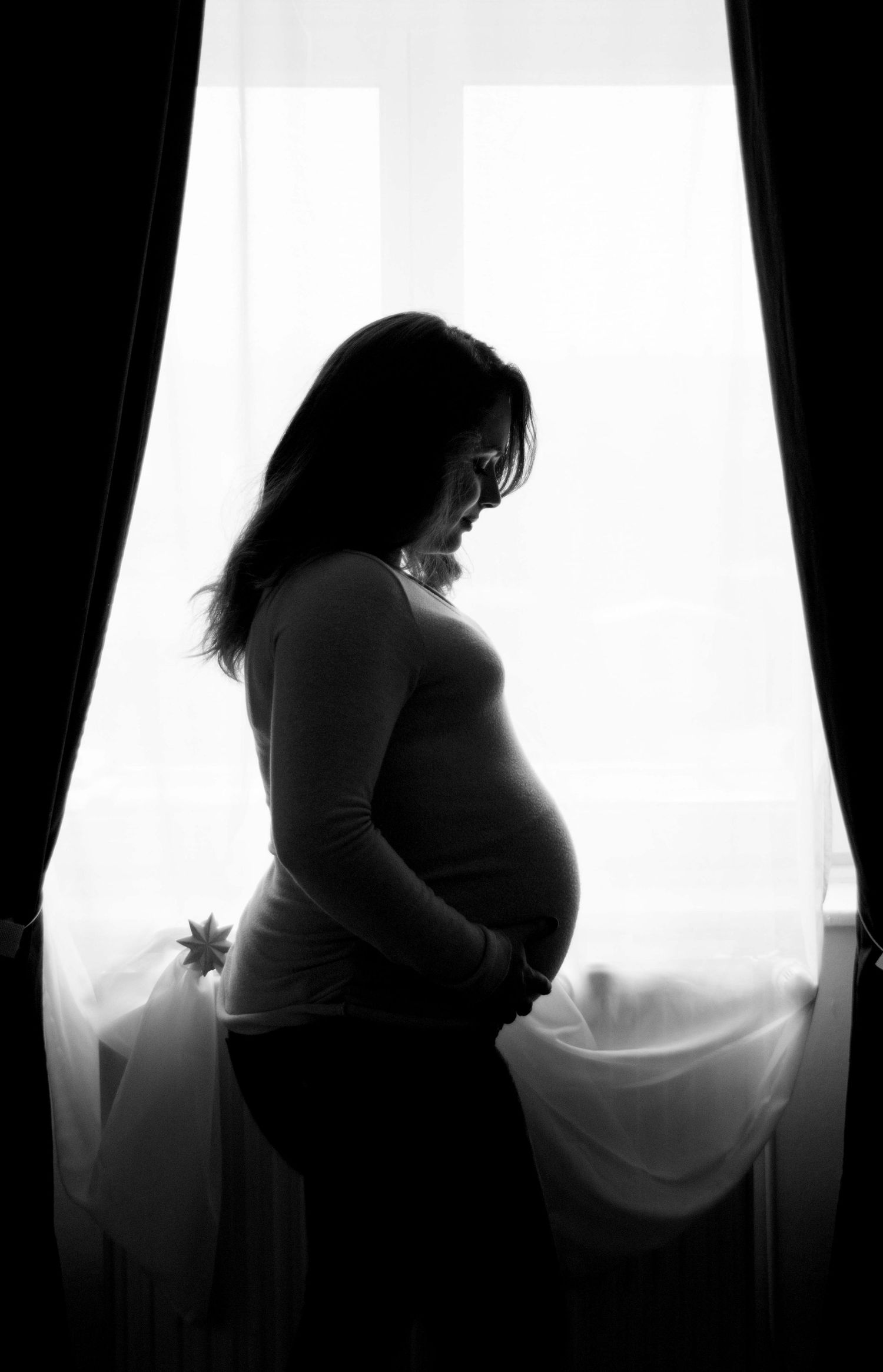 Dramatic silhouette maternity photos against bright window with dark curtains.