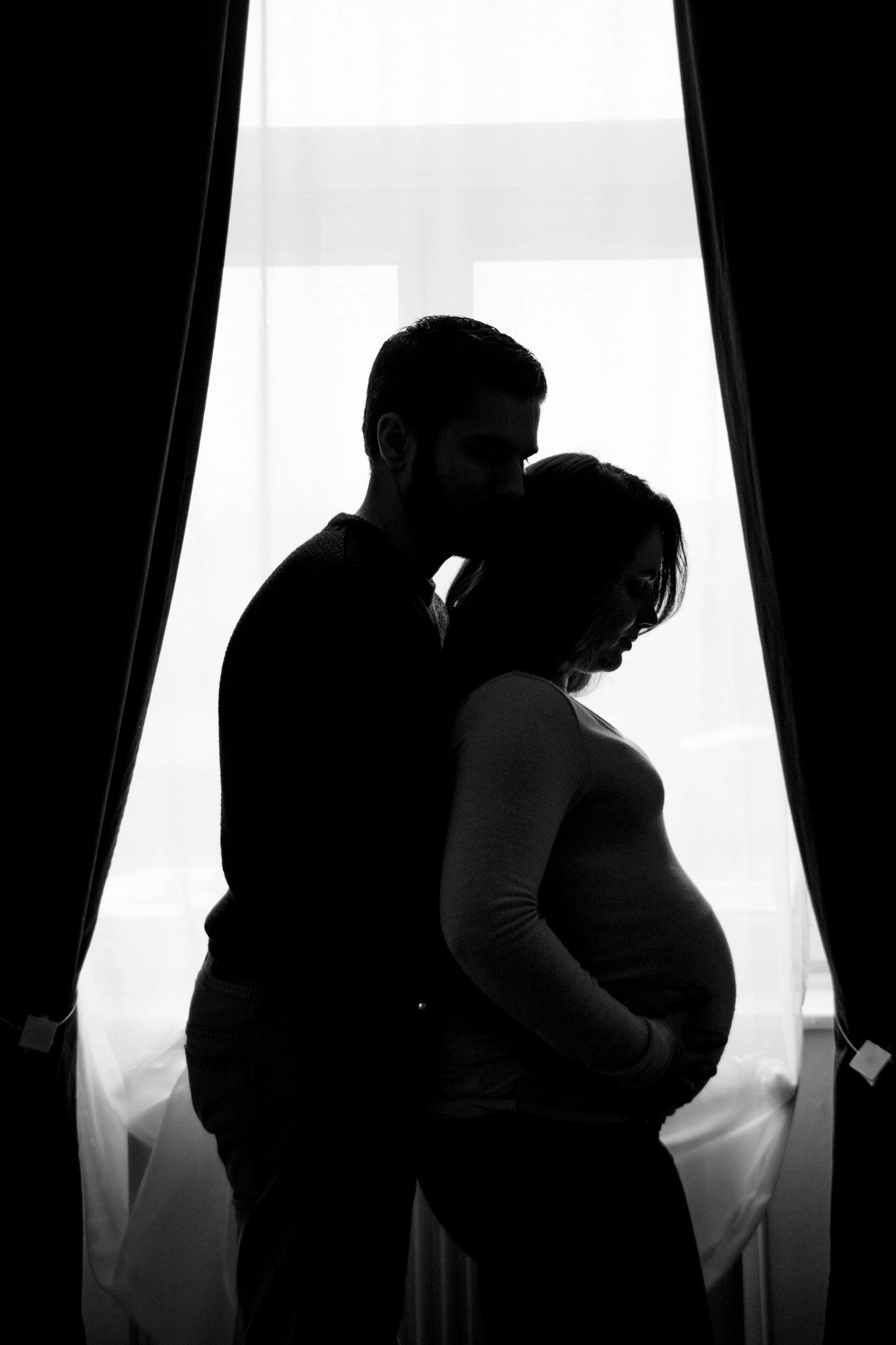 Silhouetted figures embracing by a bright window with dark curtains.
