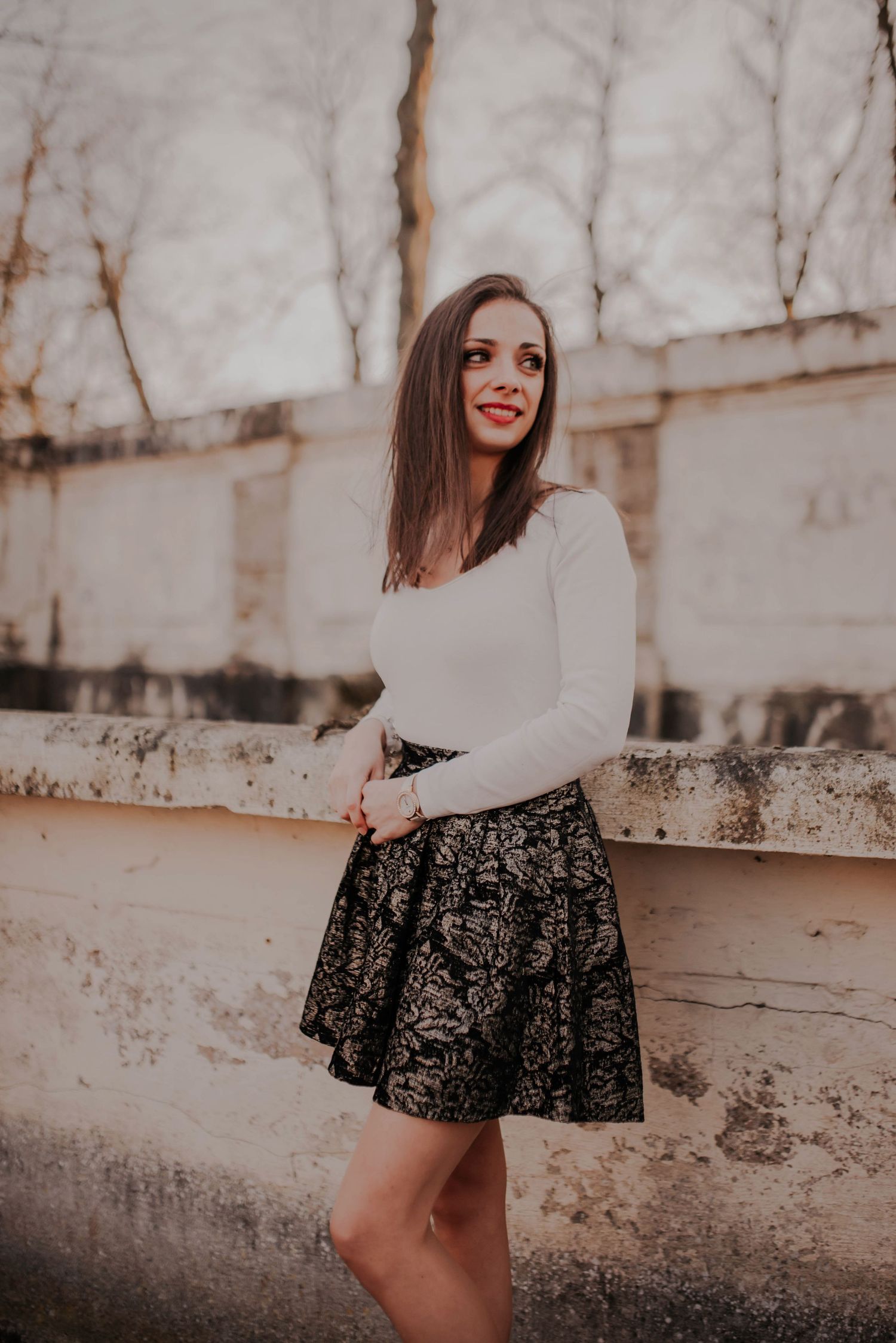 Woman in white sweater and patterned skirt poses against textured wall backdrop.