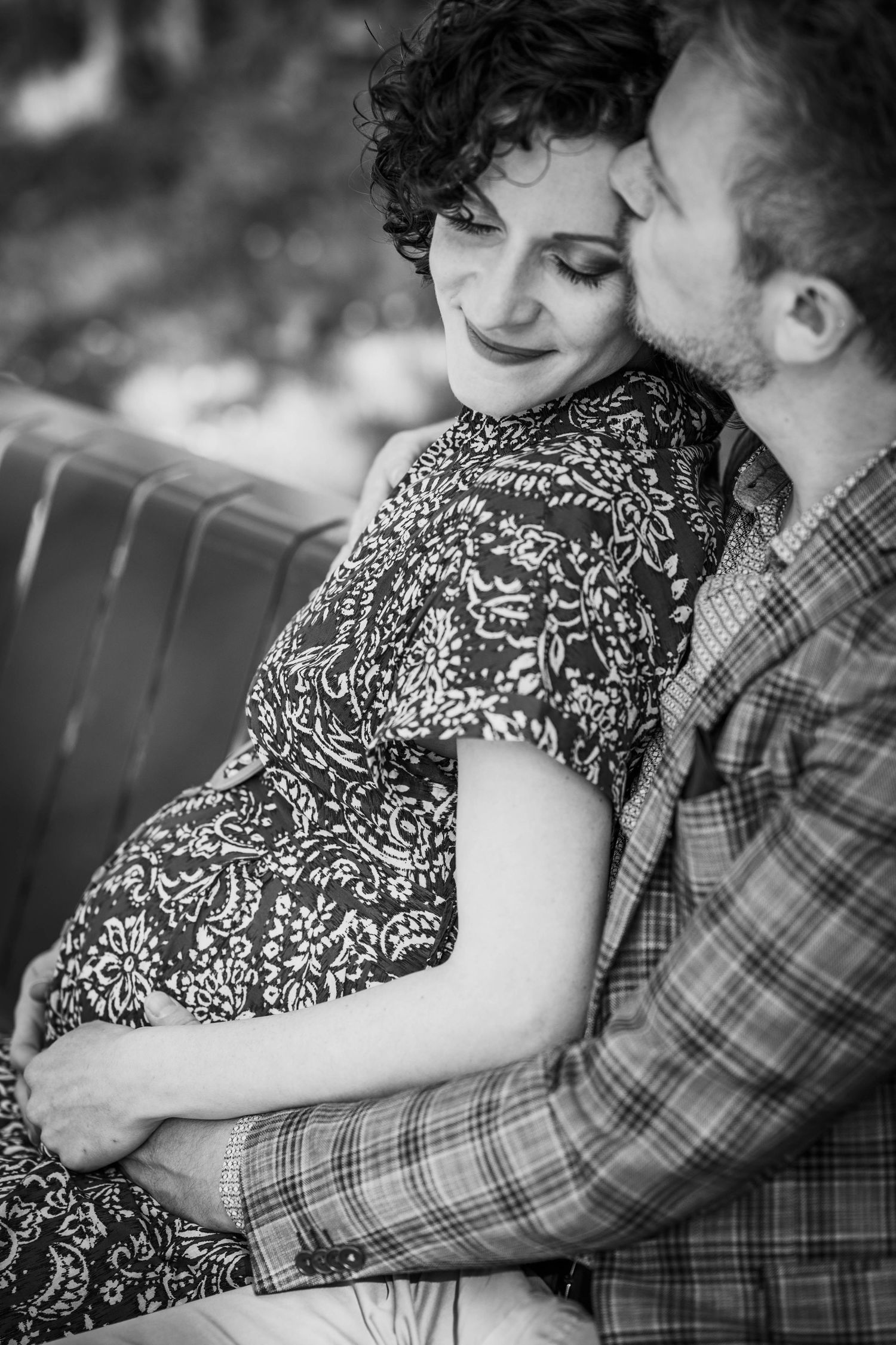 Black and white photo of a couple sitting together, man tenderly kissing pregnant woman's cheek while holding her belly.