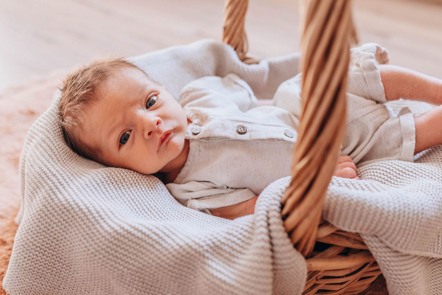 Newborn baby lying in a wicker basket wrapped in a soft beige knit blanket wearing a white onesie.