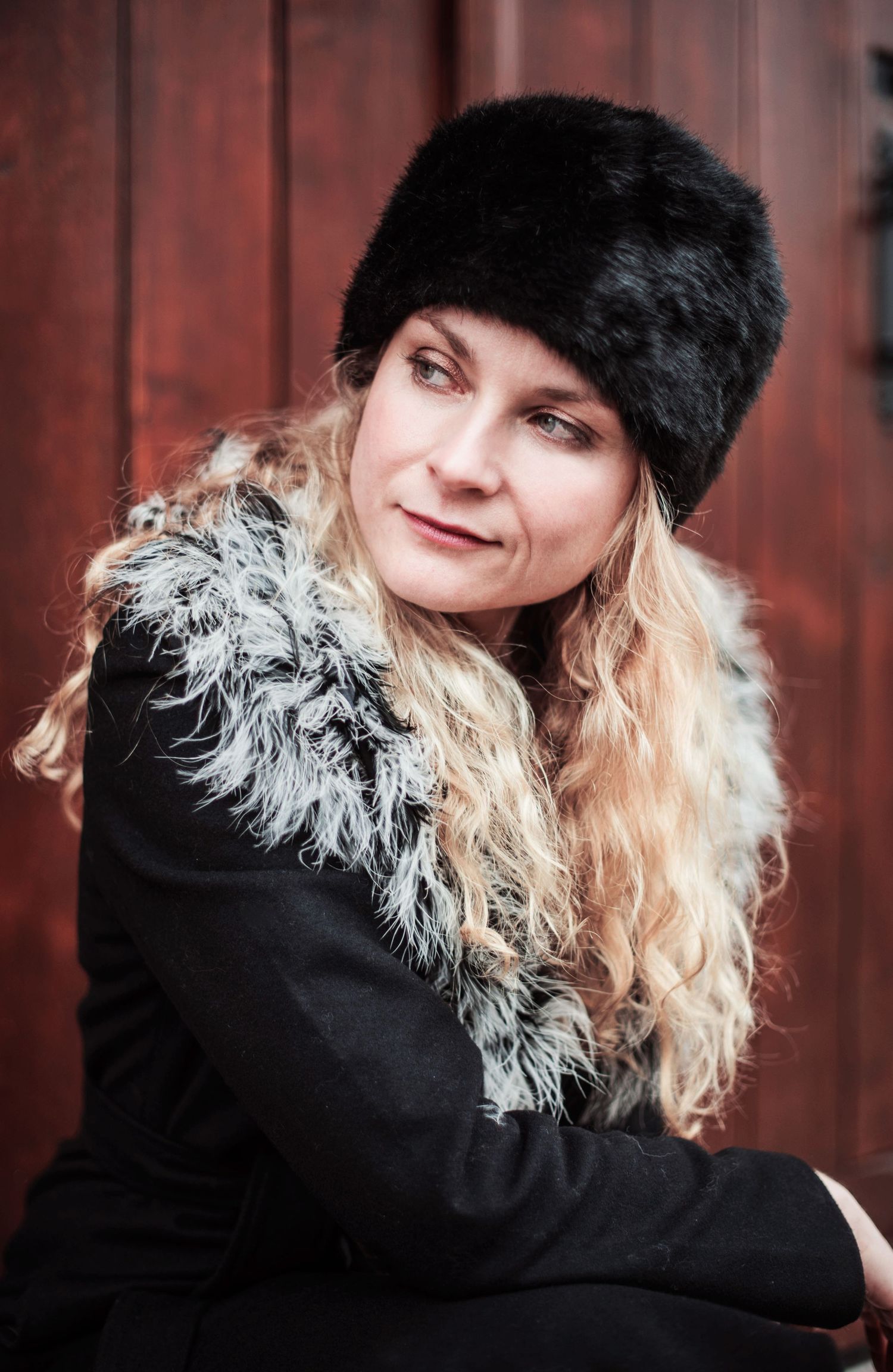 Stylish winter fashion with black fur hat and feather detail against red wooden backdrop.