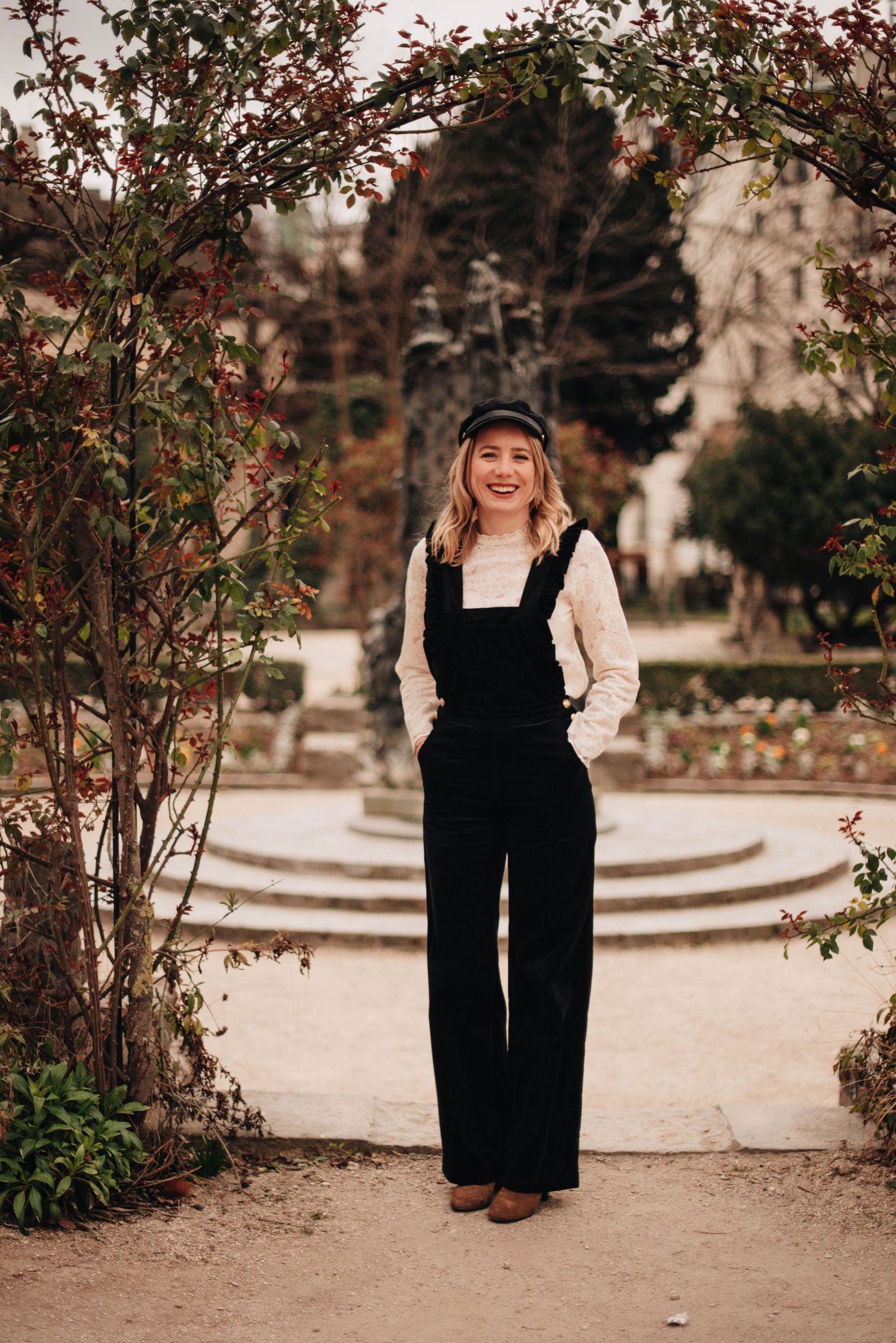 Person wearing black overalls and white top poses in a garden setting.