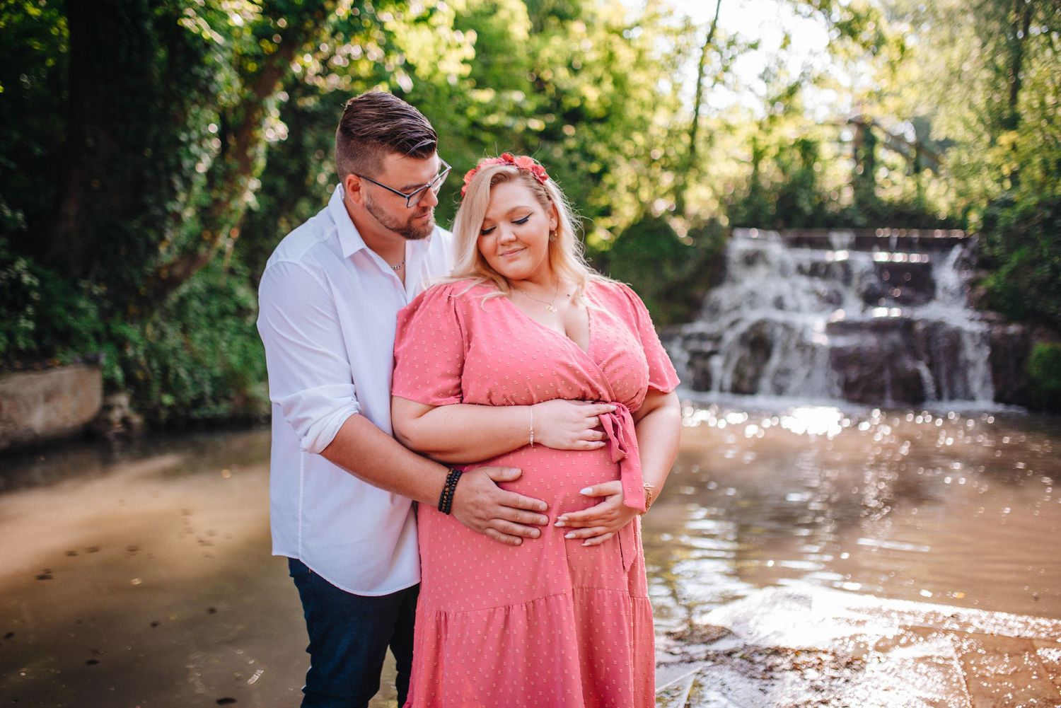 A couple poses for romantic maternity photos near a waterfall in natural lighting.