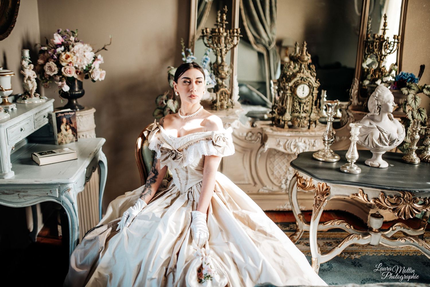 A model in an elegant pink period dress poses gracefully in a luxurious room with ornate decor and chandeliers.