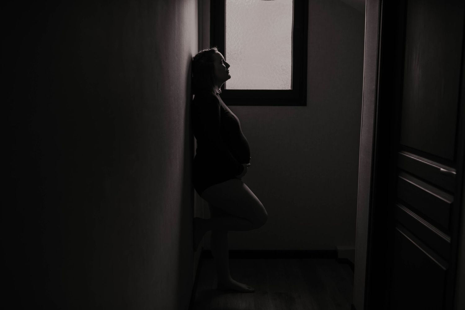 Silhouette of pregnant woman standing by window in dark hallway.