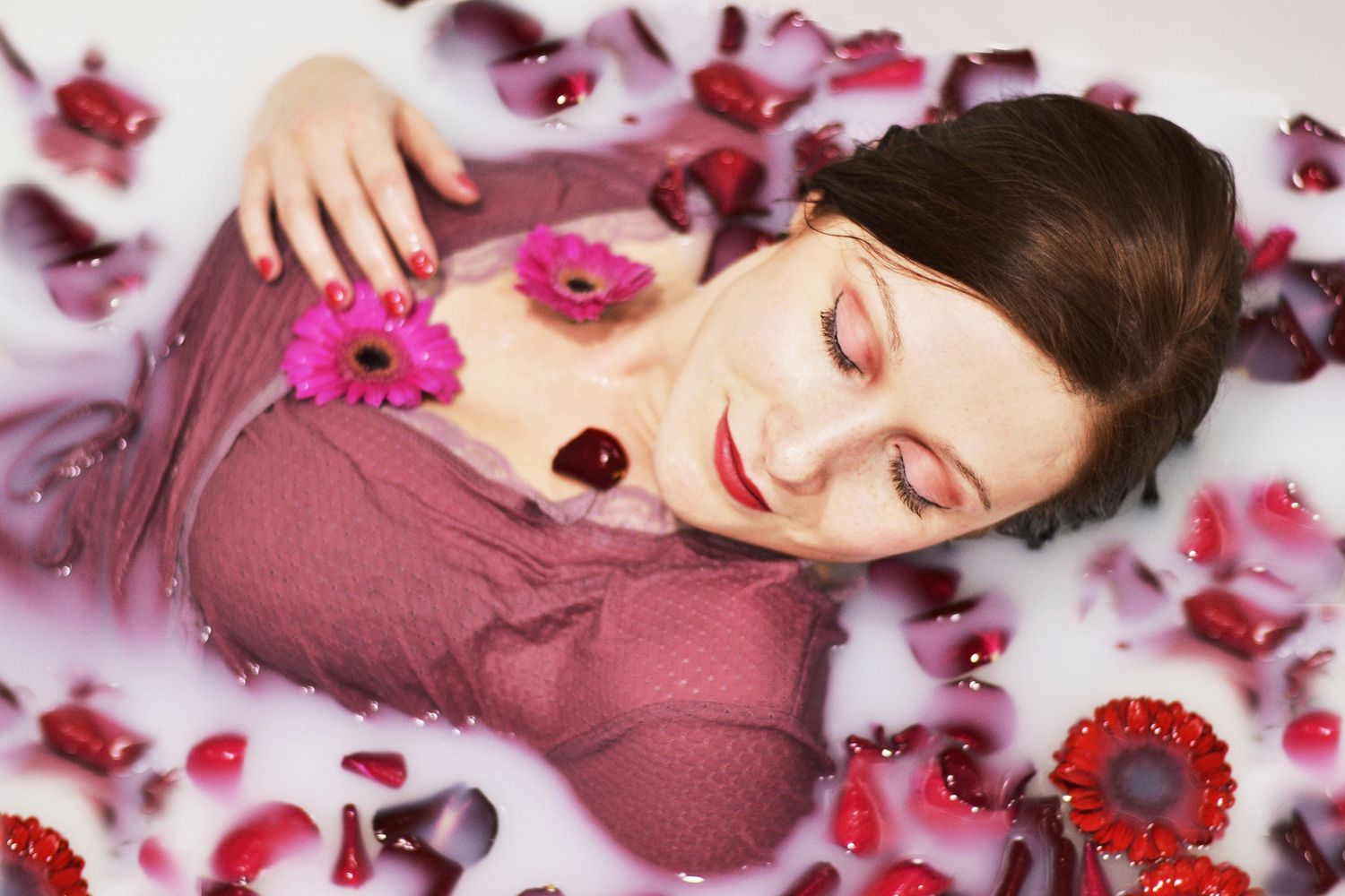 Person relaxing in bath with pink flower petals and wearing burgundy top.