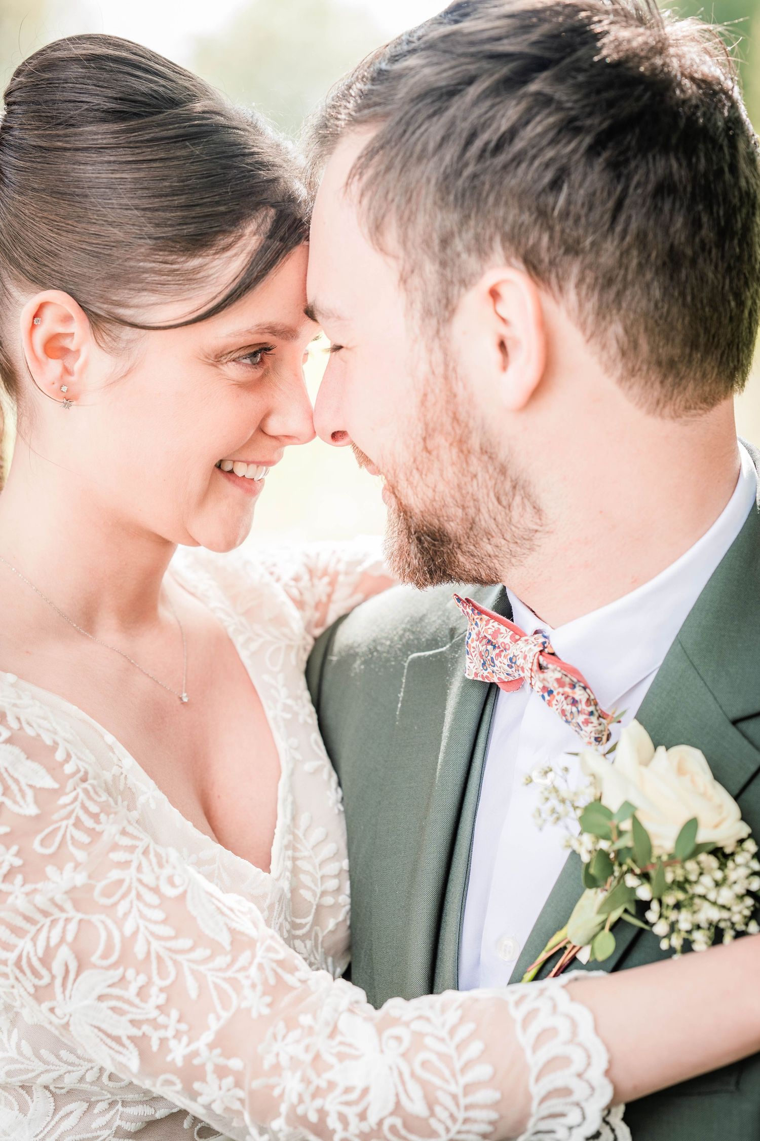 A smiling bride and groom in a green suit with floral bow tie touch foreheads tenderly on their wedding day.