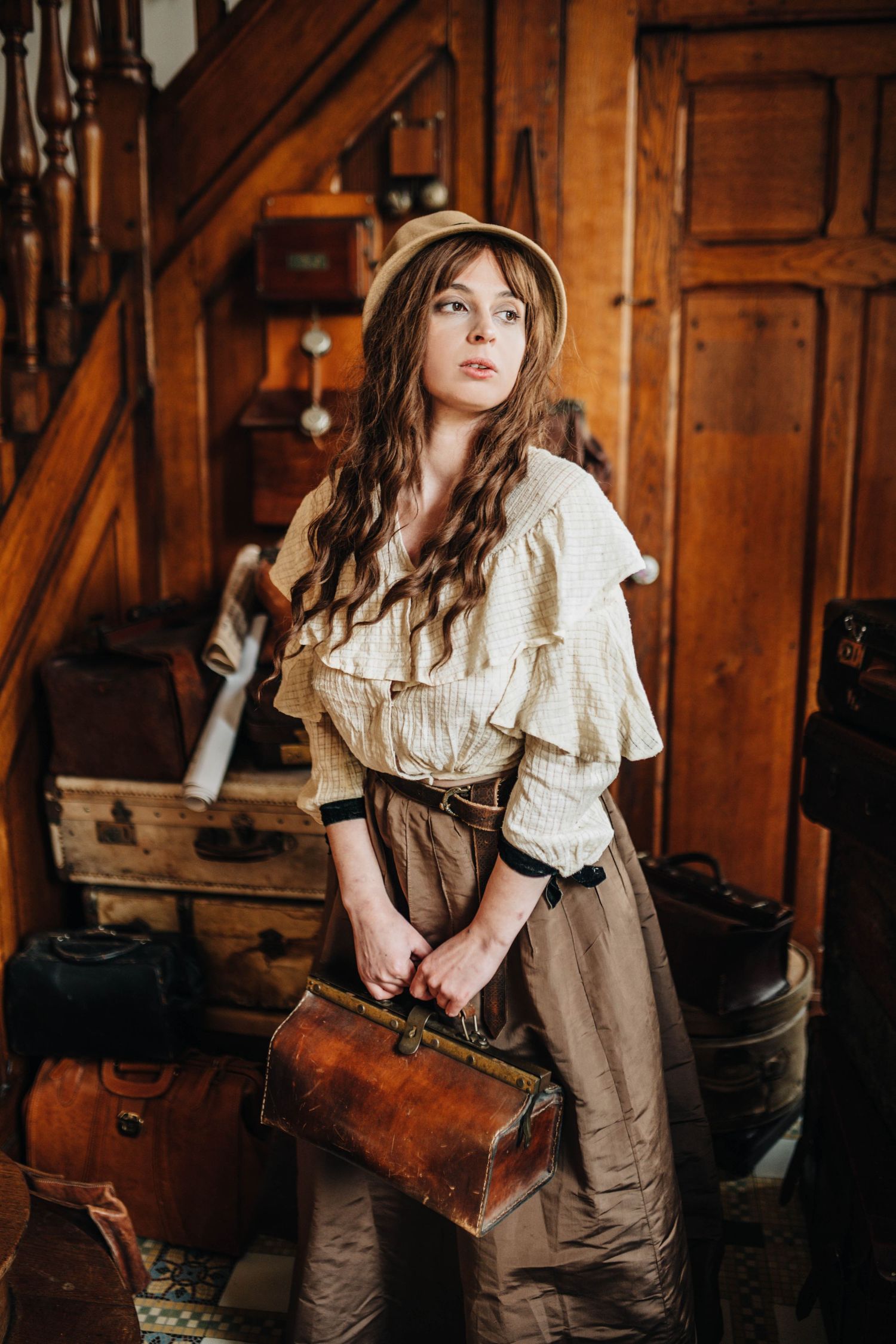 Young woman in vintage attire holding a leather bag surrounded by antique luggage in a rustic wooden interior.