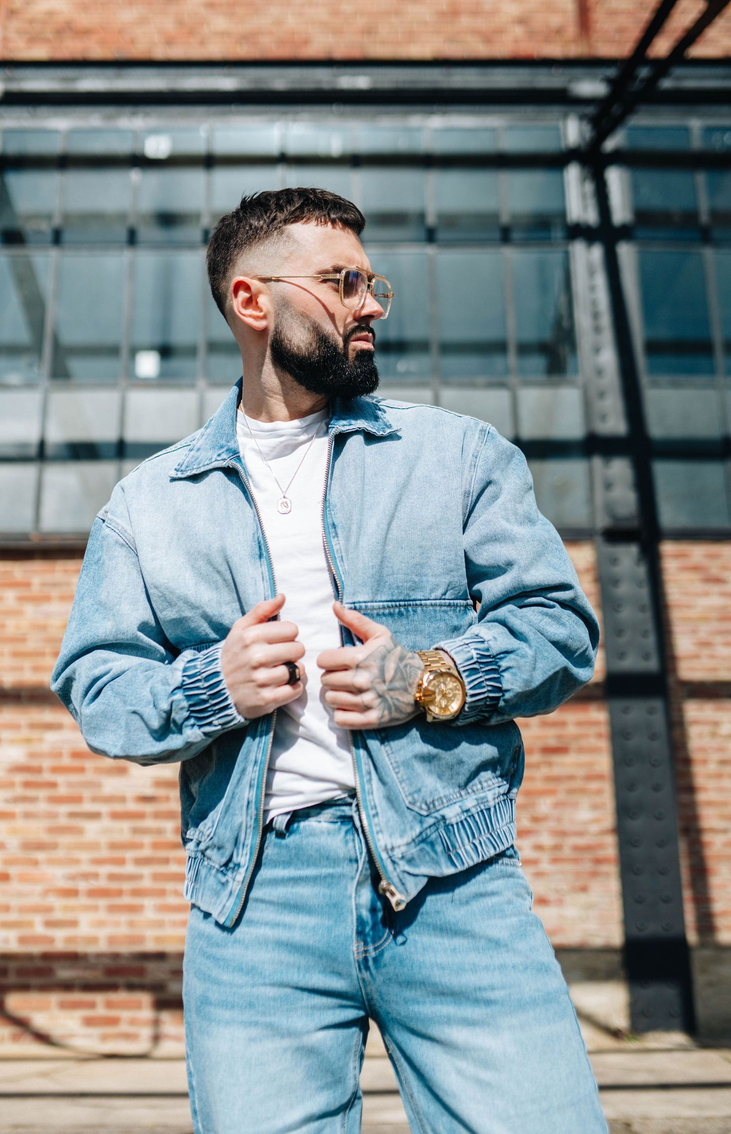 A bearded man in a denim jacket and jeans poses outdoors wearing round sunglasses and a gold watch near a brick building.