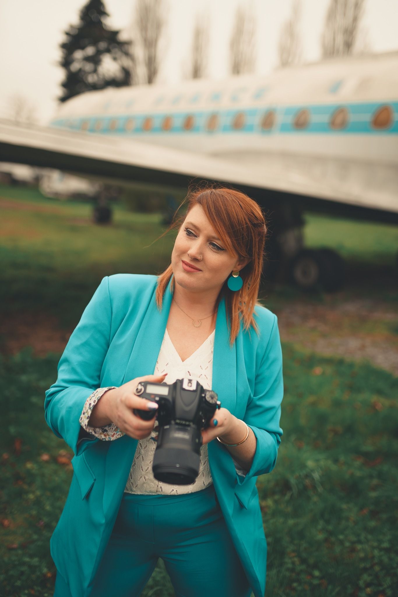 Photographer wearing turquoise blazer examines camera settings in outdoor setting.