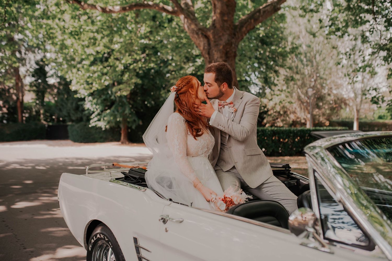 Wedding couple posing with vintage car in a sunny outdoor setting.