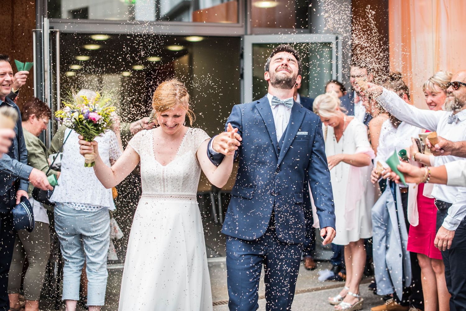 Newlyweds celebrate while guests shower them with confetti outside a building.