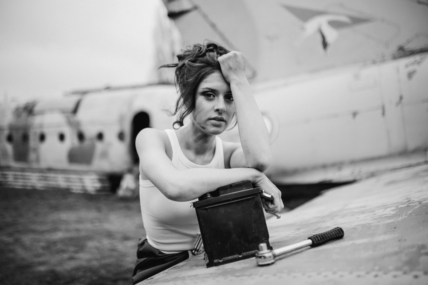 Black and white portrait of someone in a white tank top holding vintage camera equipment near an airplane.
