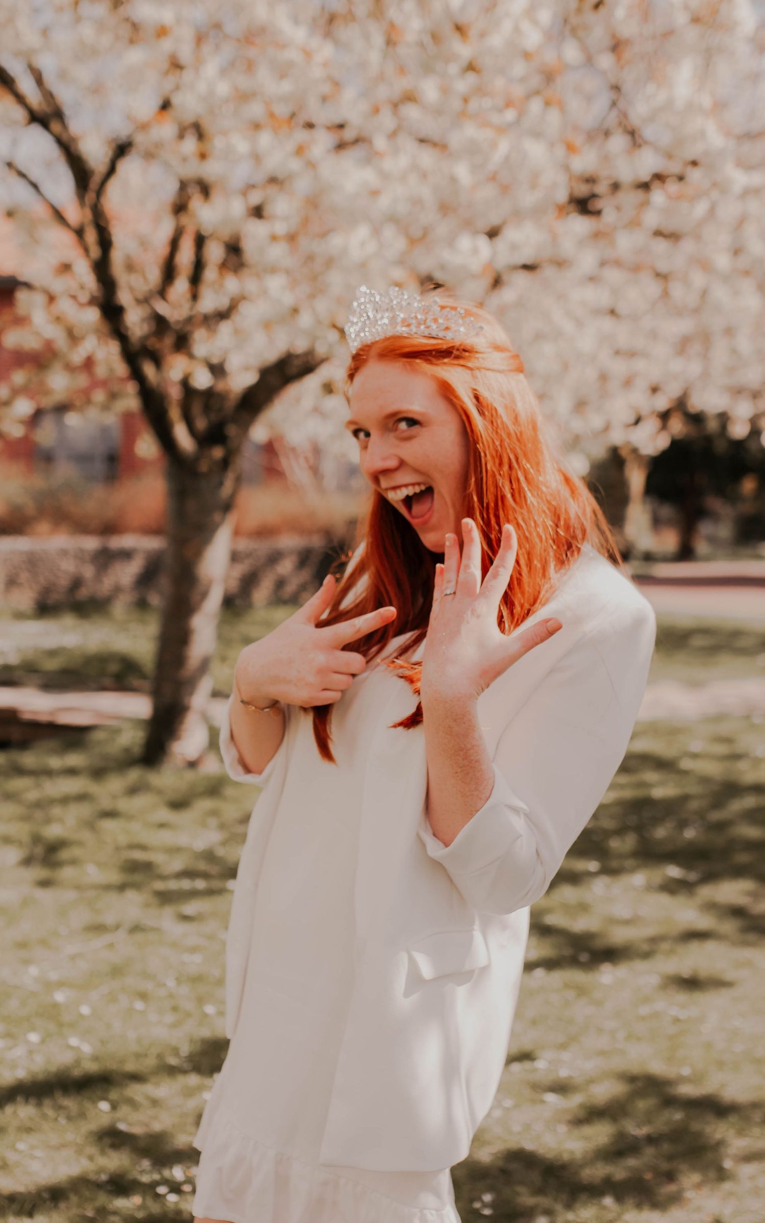 A joyful person in a white dress and tiara laughing under blooming cherry blossom trees.