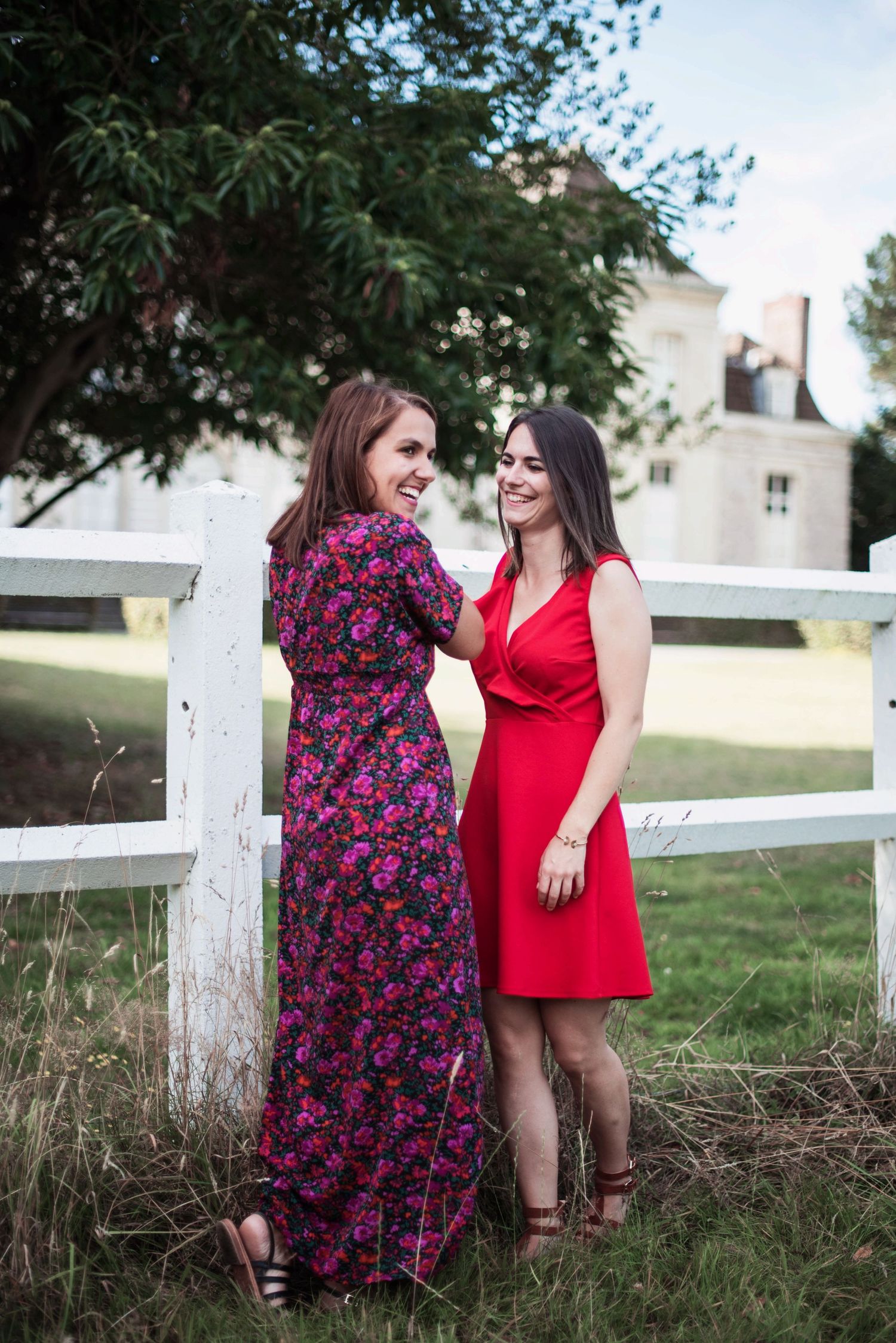 Two women in floral and red dresses pose by white fence in countryside setting.