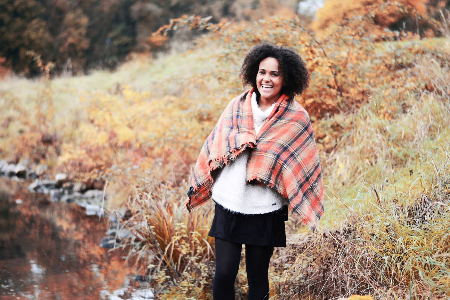 Person wearing plaid poncho smiling outdoors in autumn landscape near stream.