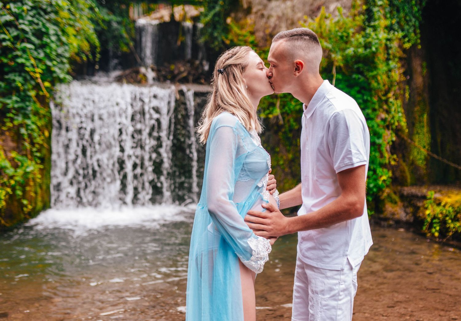 Romantic couple embracing by a scenic waterfall in matching white and blue outfits.