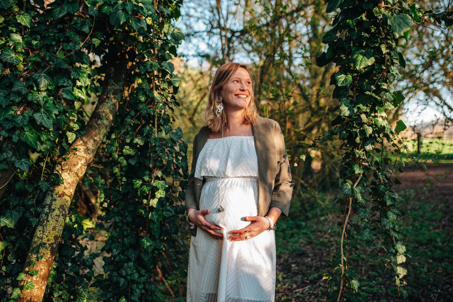 Maternity photoshoot in a white dress against lush green foliage with natural sunlight filtering through.