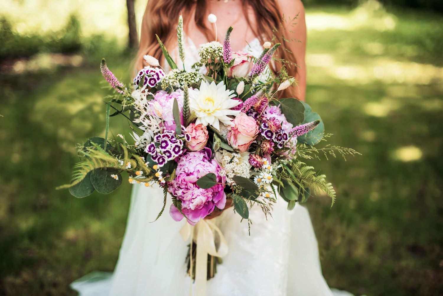 Stunning bridal bouquet features pink and purple flowers with delicate greenery against sunlit background.