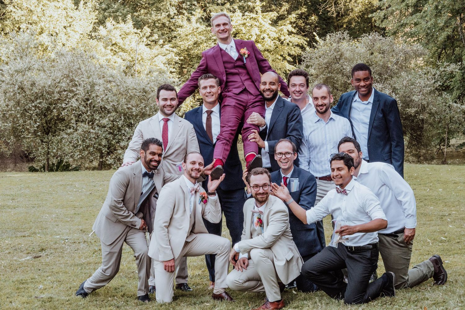 Group of groomsmen in suits posing outdoors, lifting groom in burgundy suit during wedding celebration.