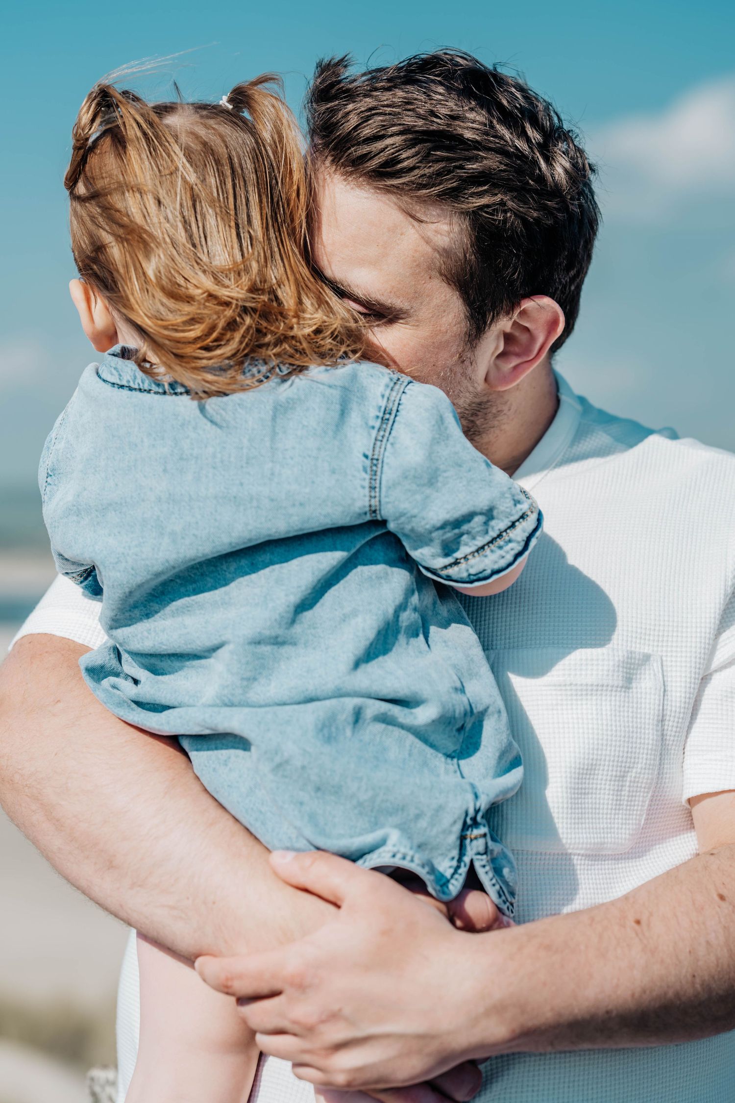 Father kissing young child in denim outfit outdoors under blue sky, tender parenting moment.