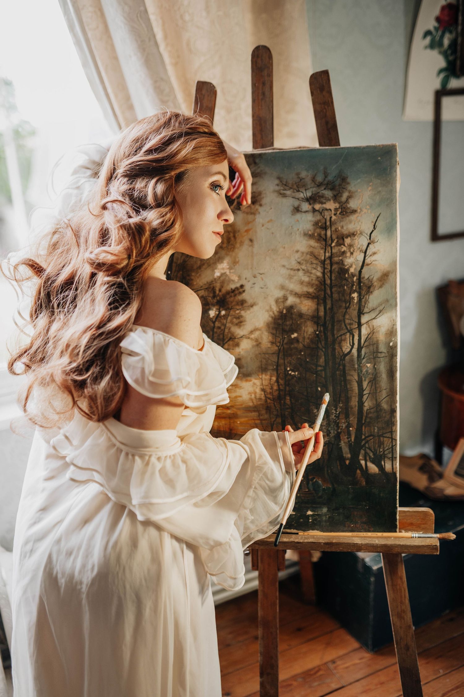 A woman in a white off-shoulder dress painting a forest landscape on an easel in a sunlit studio.