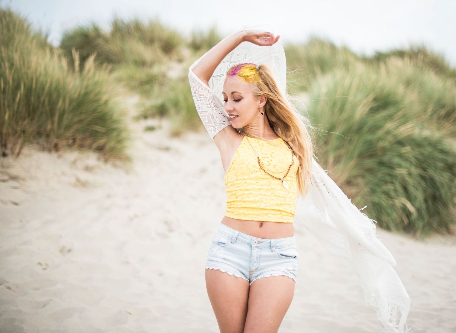 A summer beach scene with someone in casual wear posing among coastal grass and sand dunes.