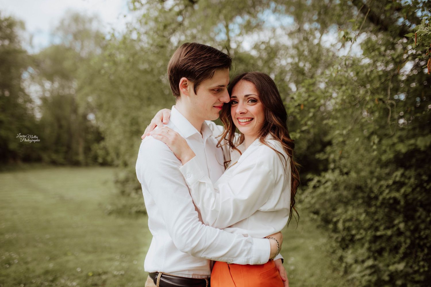 Romantic outdoor photoshoot with couple in white and orange outfits against natural backdrop.