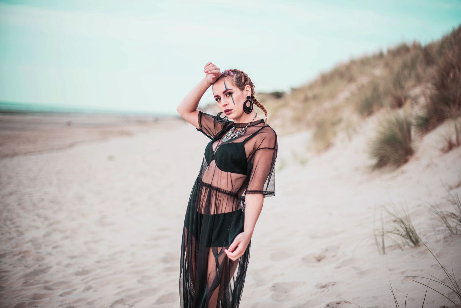 Model in black sheer dress poses dramatically on sandy beach with moody lighting.