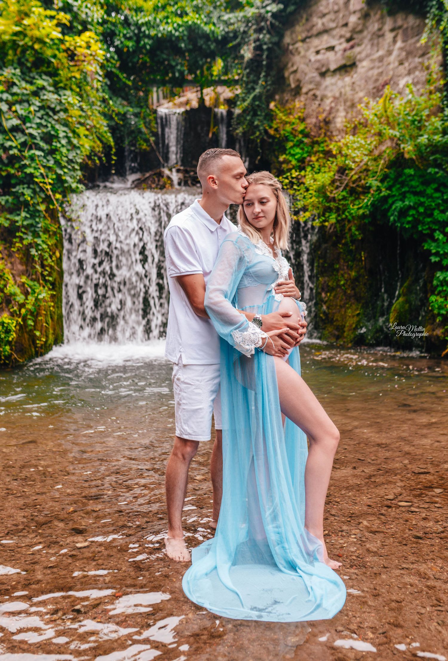 A romantic maternity photoshoot at a waterfall with a couple in white and light blue outfits.