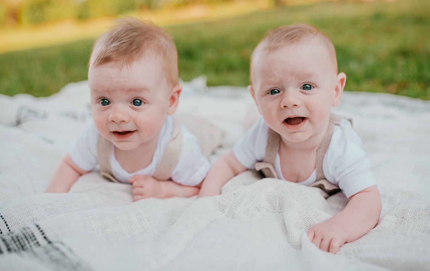 Twins smiling while lying on white blanket during outdoor portrait session.