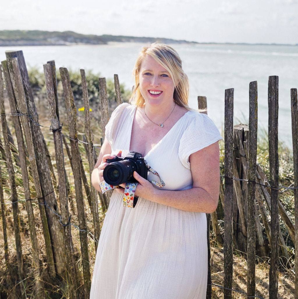 Photographer with camera posing near wooden beach fence during seaside photoshoot.