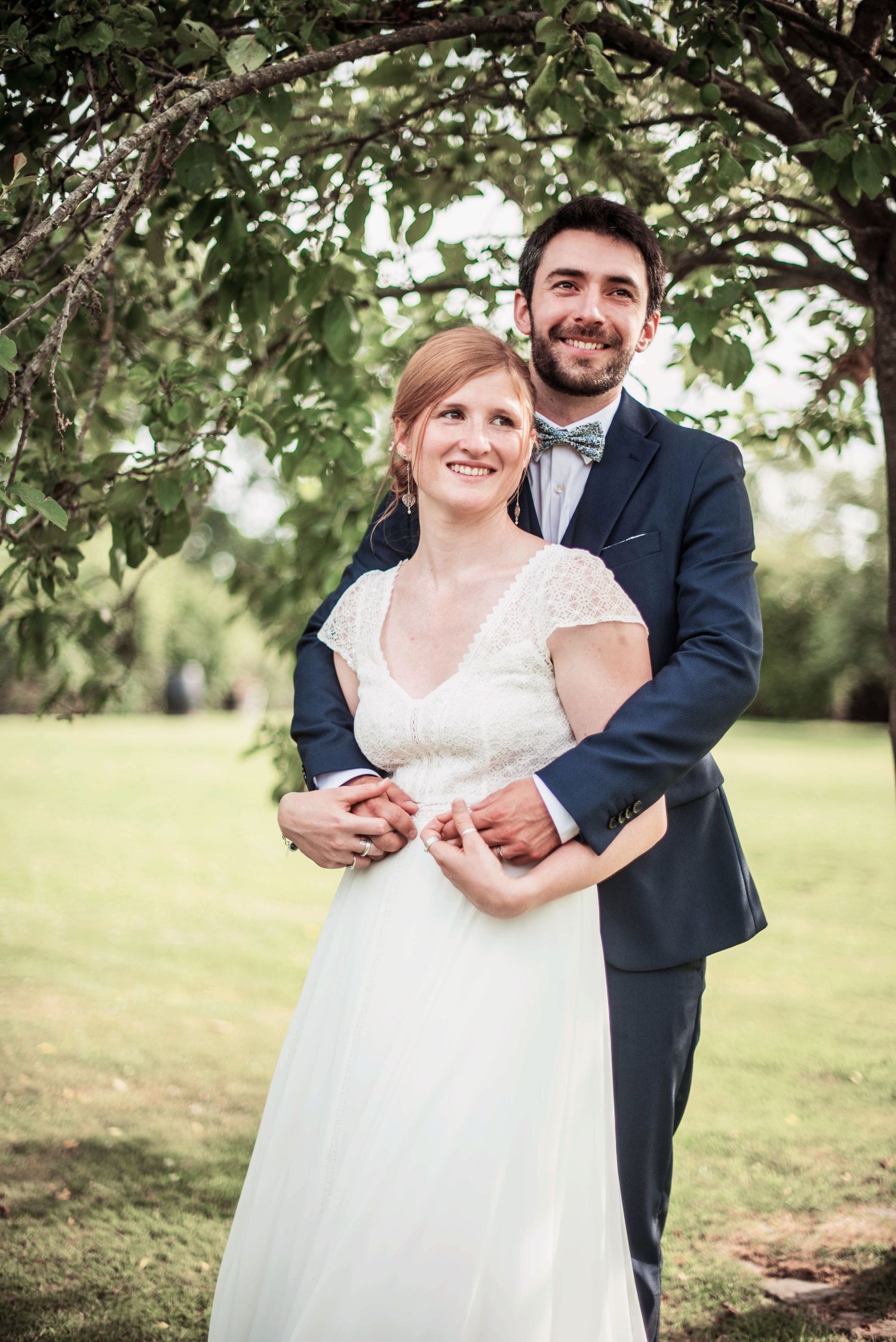 A couple shares an intimate embrace under green leafy trees in their wedding attire.