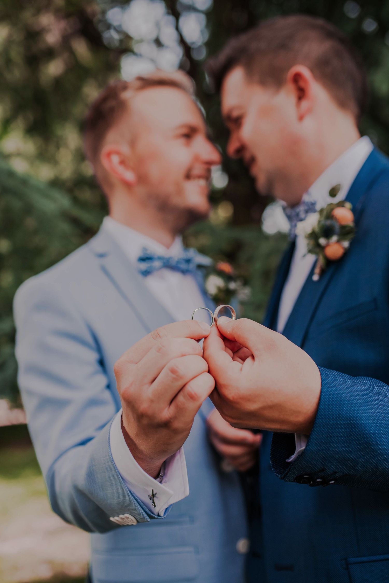 Two people in suits share an intimate moment exchanging rings during an outdoor wedding ceremony.