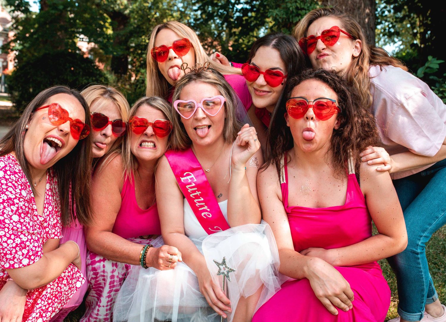 A bachelorette party group wearing pink heart-shaped sunglasses poses together for a fun photo.
