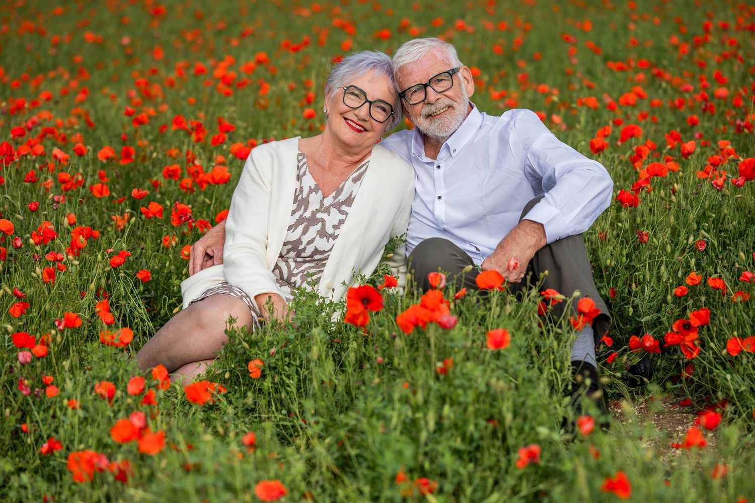 Happy senior couple sitting together in a vibrant red poppy field, smiling warmly on a sunny day.