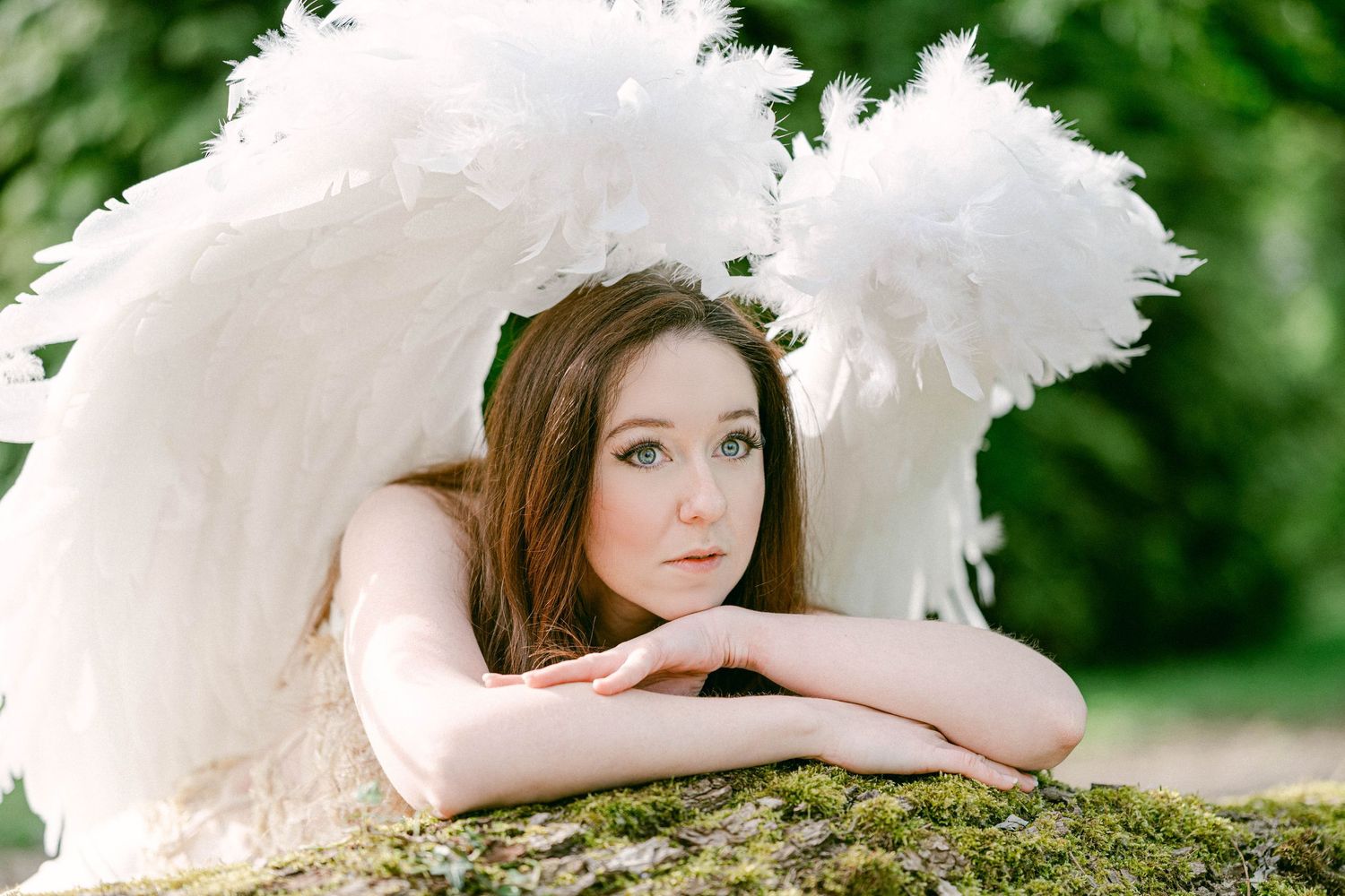 Young woman with angel wings costume lying on mossy rock in garden, looking dreamily at camera.
