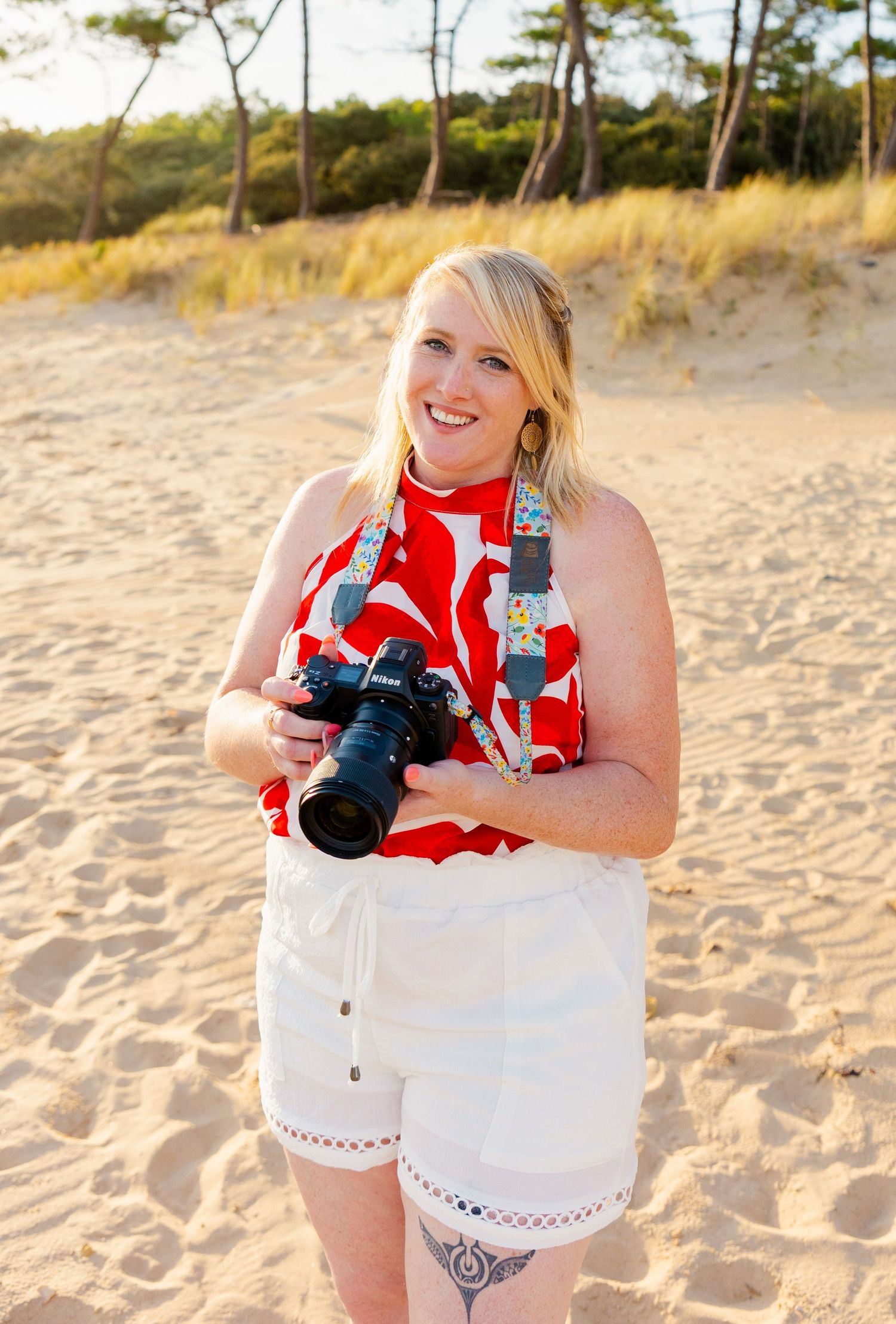 Photographer in white outfit holding cameras on sunny beach with sand and trees in background.