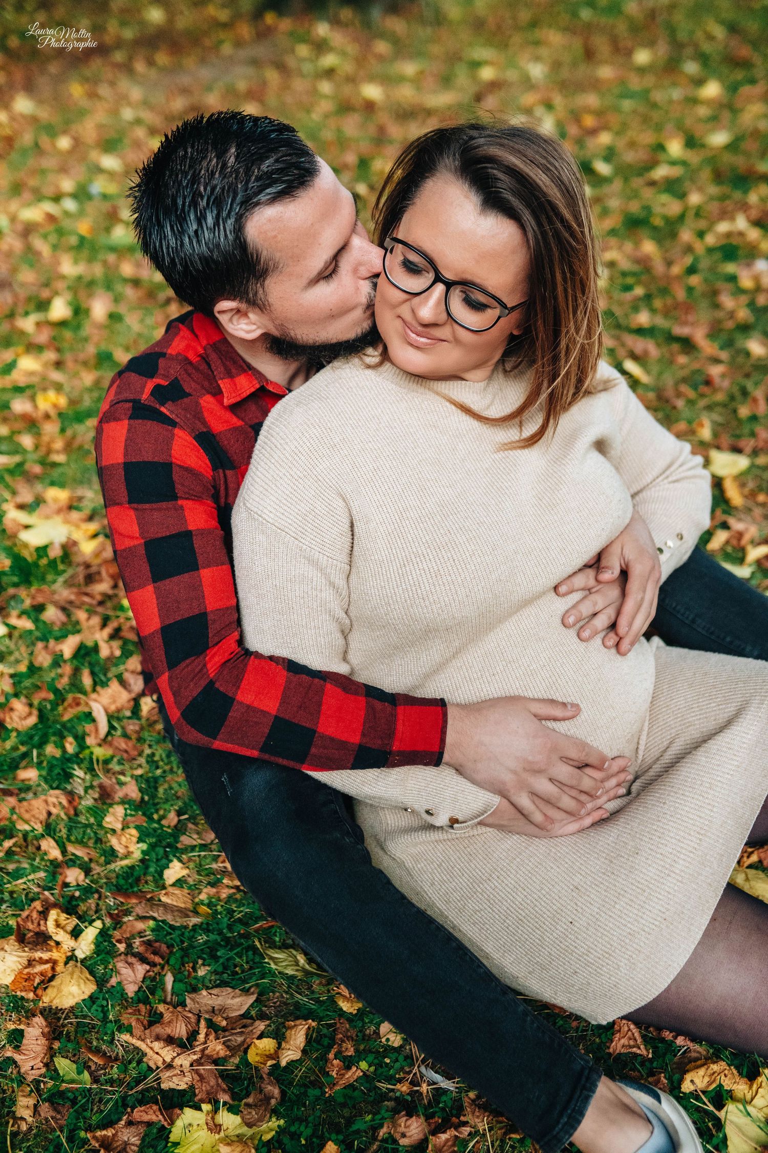 Autumn maternity photo with couple in plaid and cream sweater among fallen leaves.