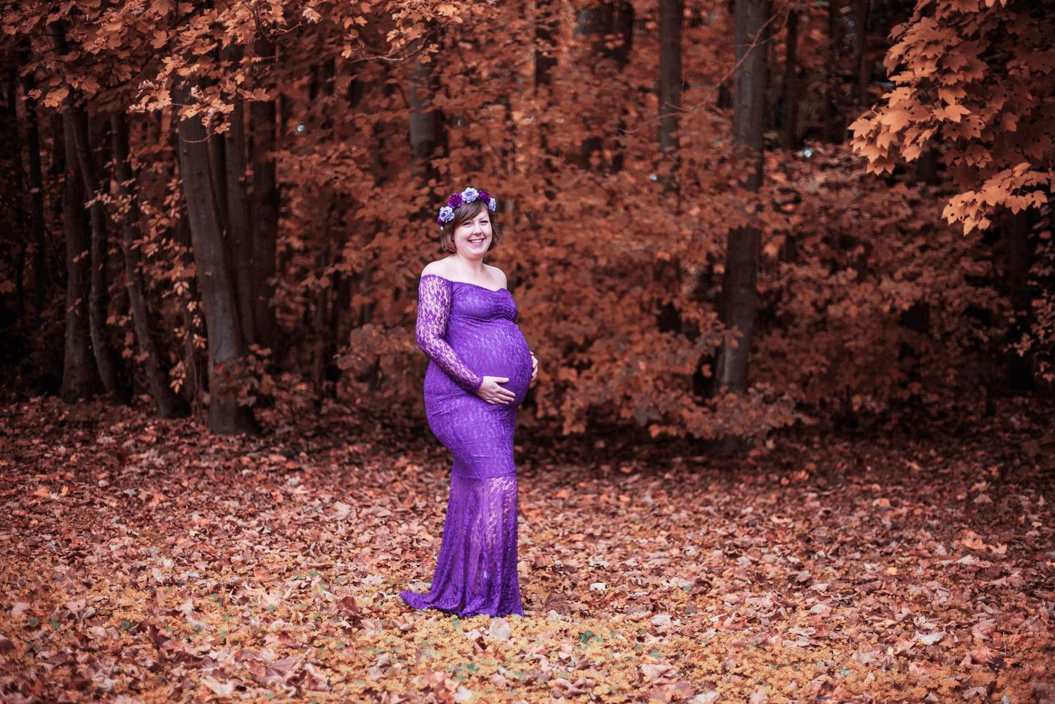 Maternity portrait in a purple gown against an autumn forest backdrop with orange-tinted leaves.