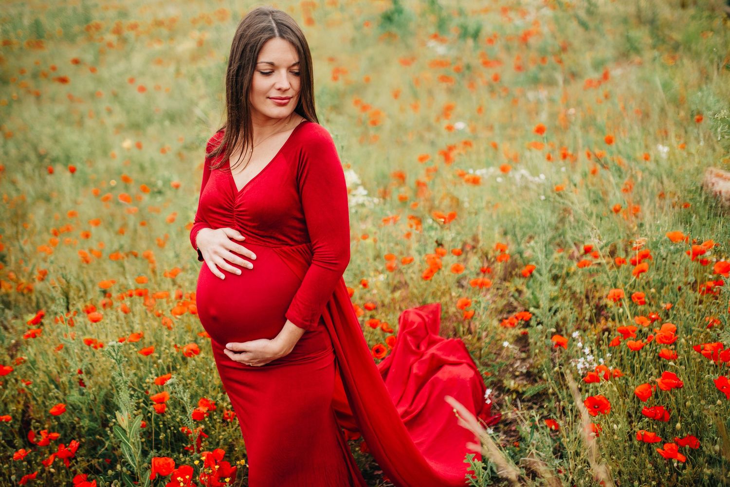 Maternity portraits in a vibrant red dress among blooming poppies in a field.