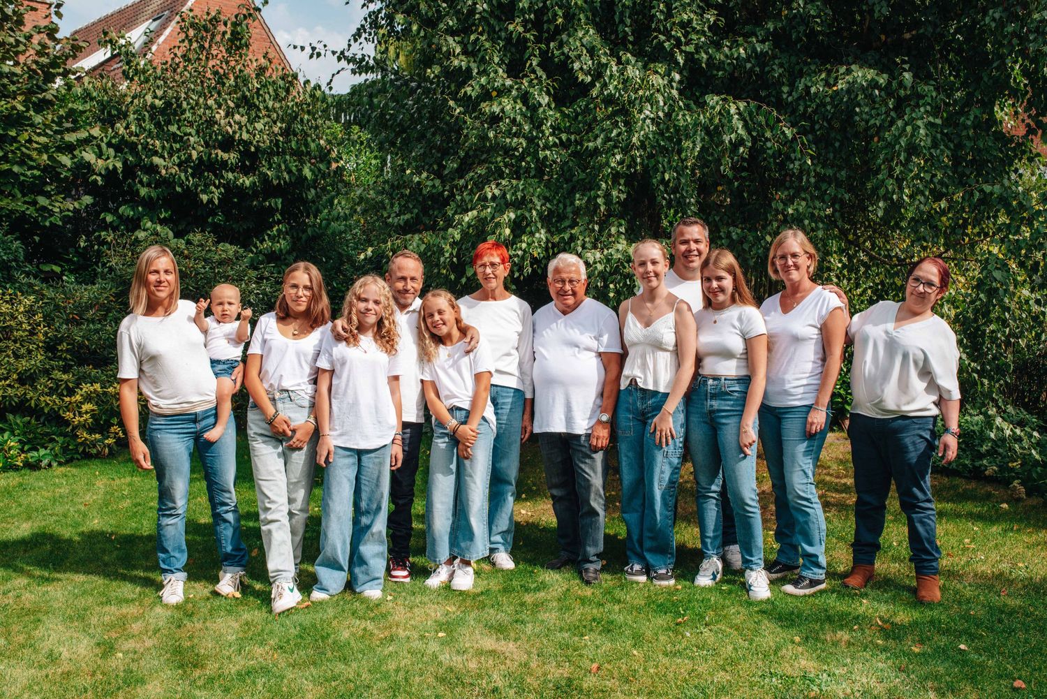 A large group poses outdoors wearing matching white shirts and blue jeans in a garden setting.
