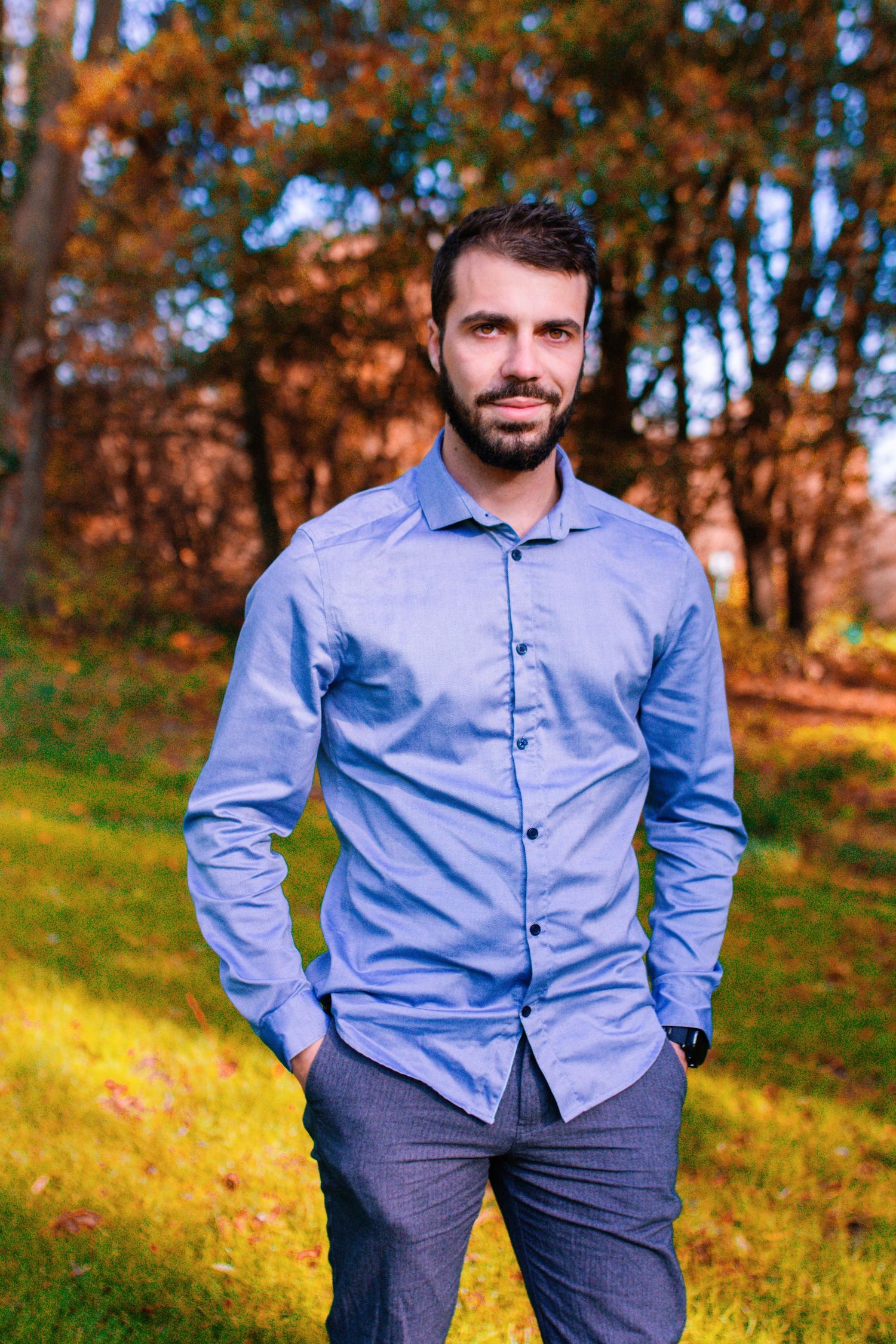Man in light blue button-down shirt poses in autumn park setting.