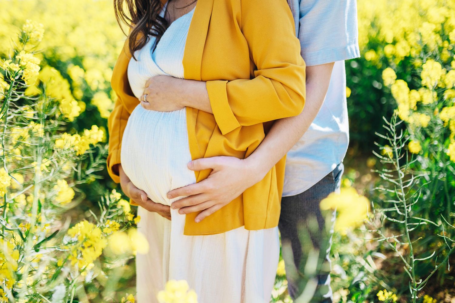 Pregnant couple embracing in yellow flower field, woman in mustard cardigan cradling baby bump outdoors.