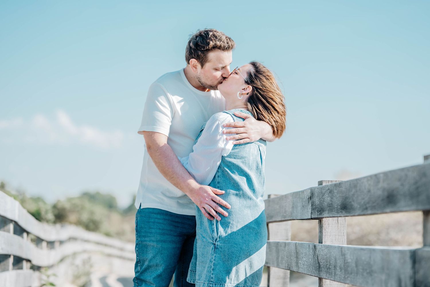 Couple sharing a romantic kiss outdoors on a wooden bridge under a clear blue sky on a sunny day.